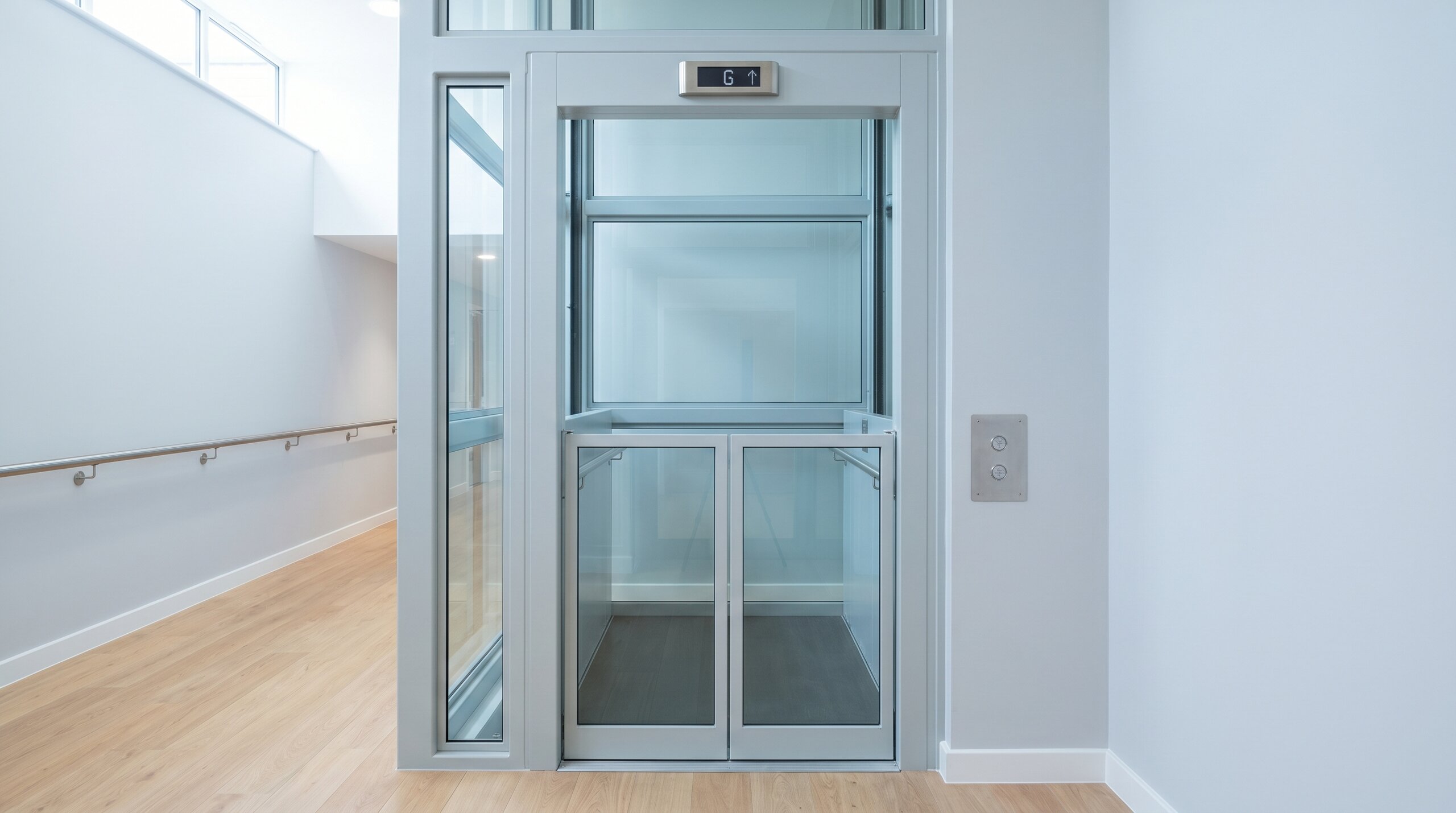 Wheelchair-accessible platform lift in glass and painted steel inside the bright foyer of a UK care home, doors closed at the lower landing with a single hall lantern above.