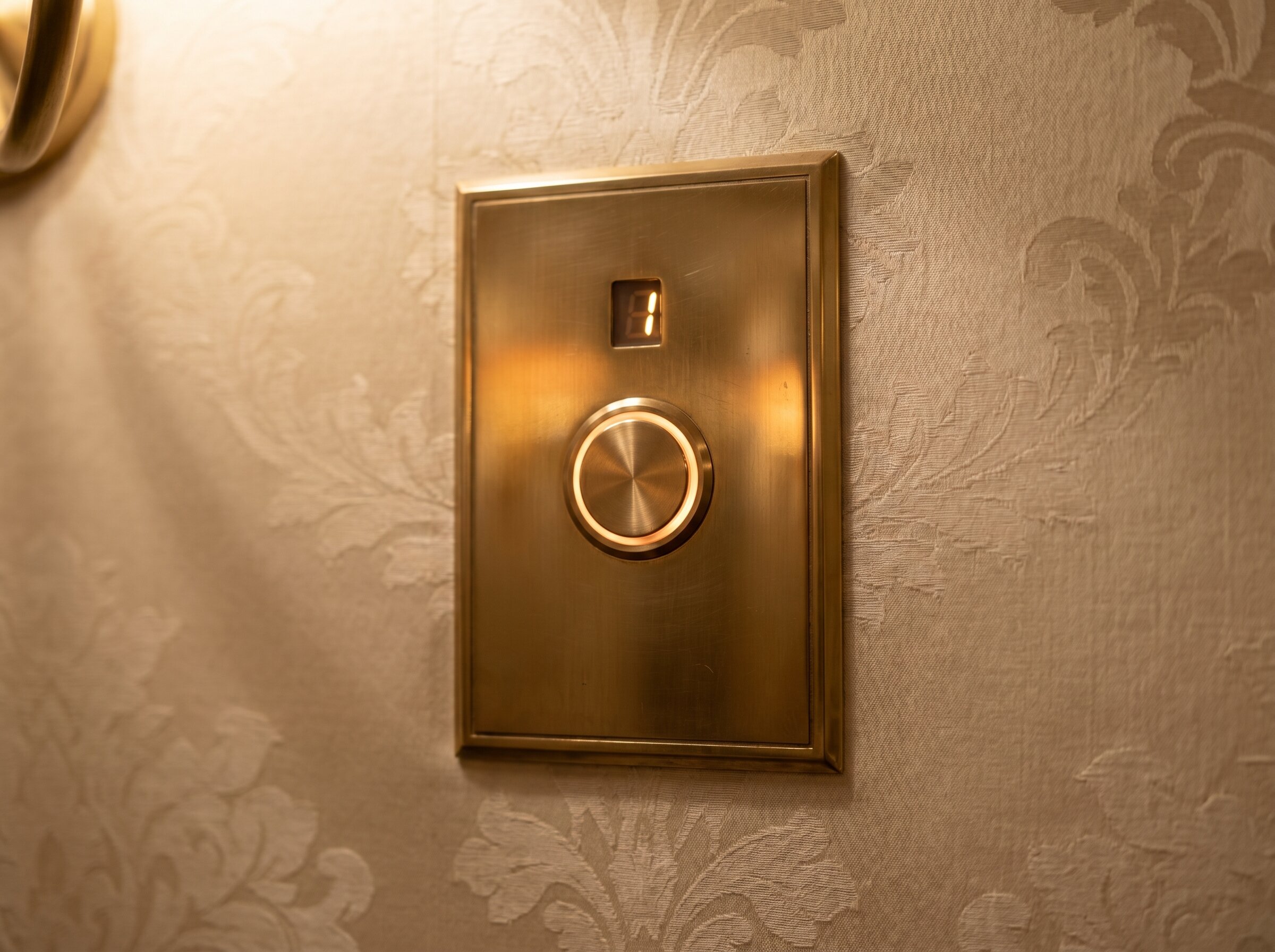 Brushed-brass round call button on an engraved brass plate beside a silk-printed wallpaper, with a small floor-indicator LED above.